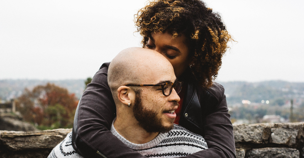 Woman hugging and kissing man's head affectionately outdoors with a scenic background.