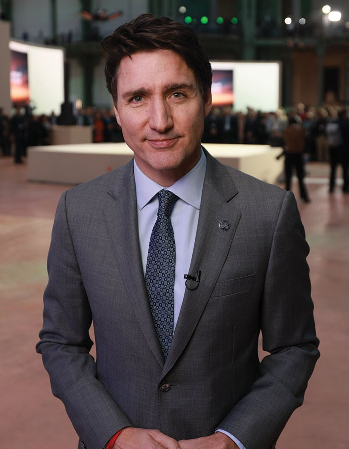 A man in a gray suit and tie stands indoors, facing the camera, with people and large screens in the background.