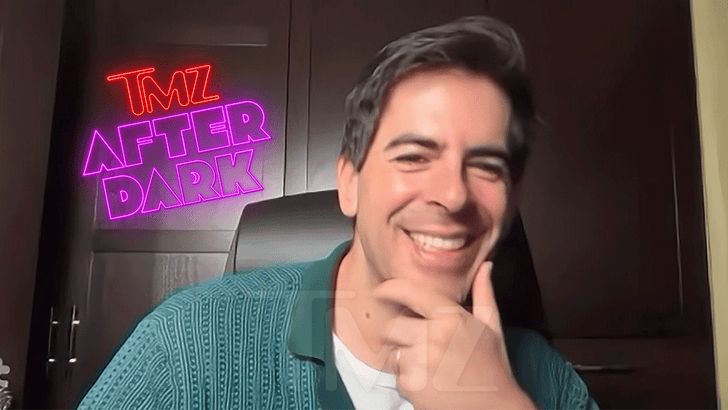 Smiling man in a green shirt on TMZ After Dark set, against a dark backdrop with neon signs.