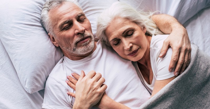 Elderly couple peacefully sleeping together, embracing under a blanket on white pillows.