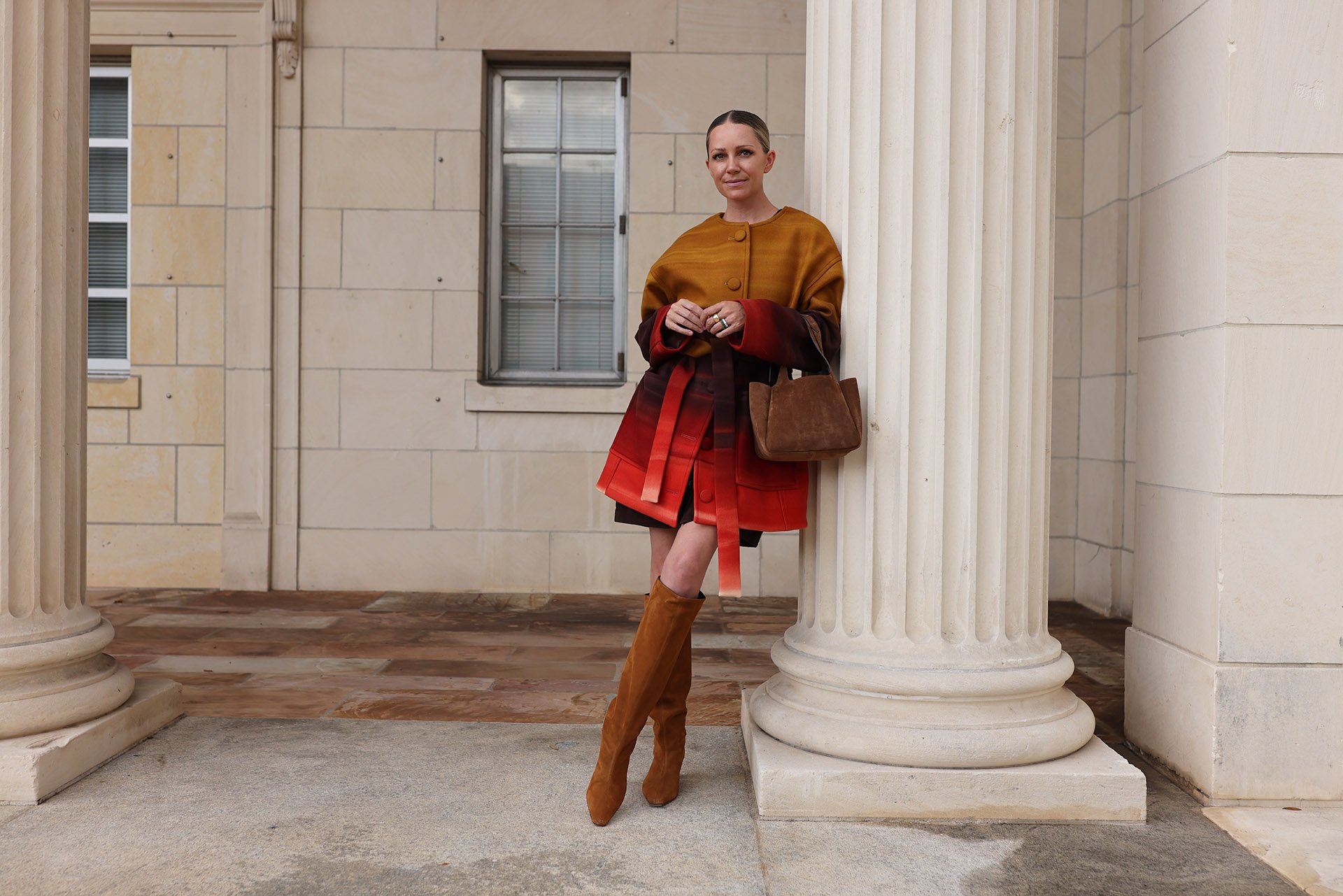 Stylish person in a colorful coat and boots leans against a column of a classic building, holding a brown bag.