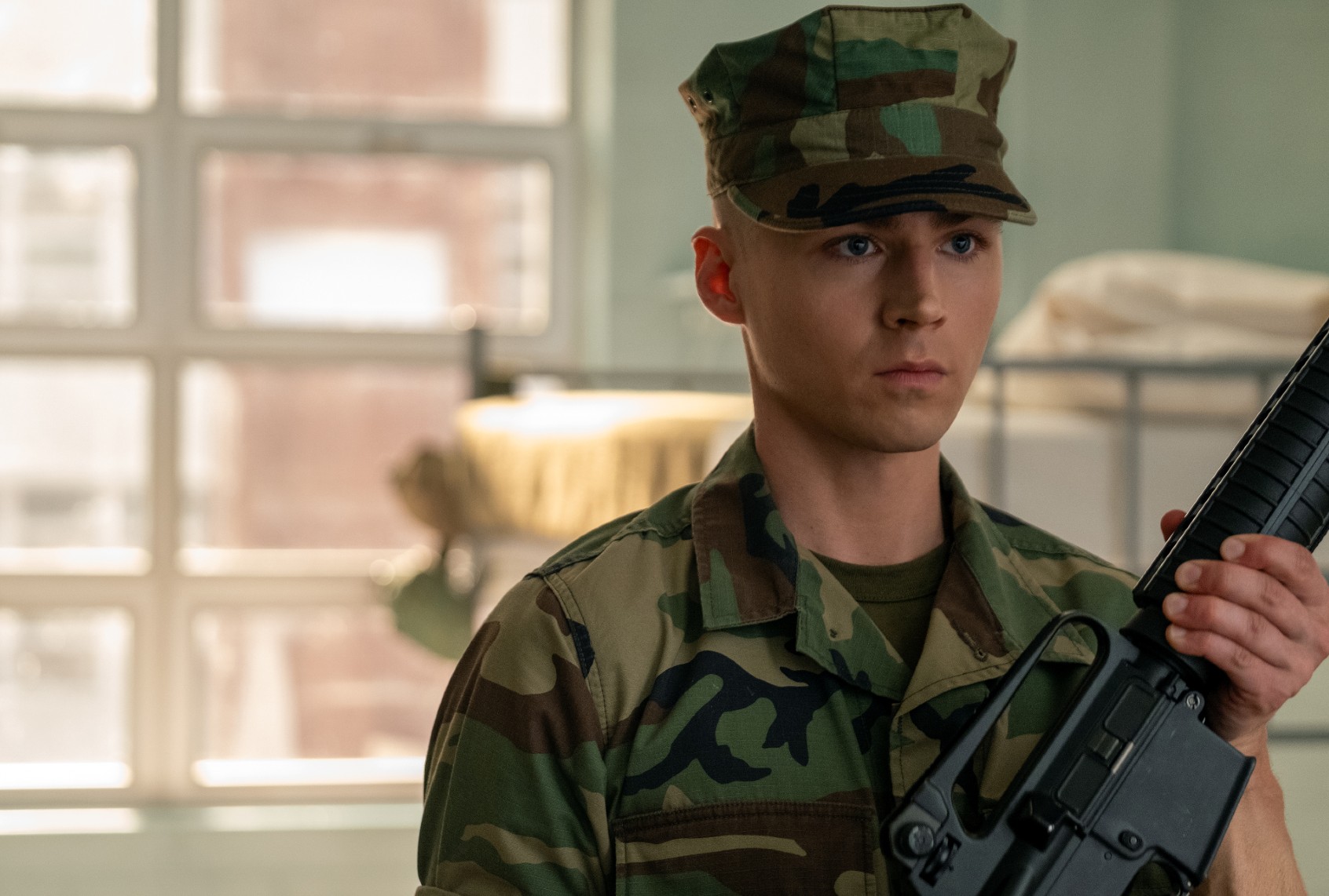 Young soldier in camouflage uniform holds a rifle, standing indoors by a window.
