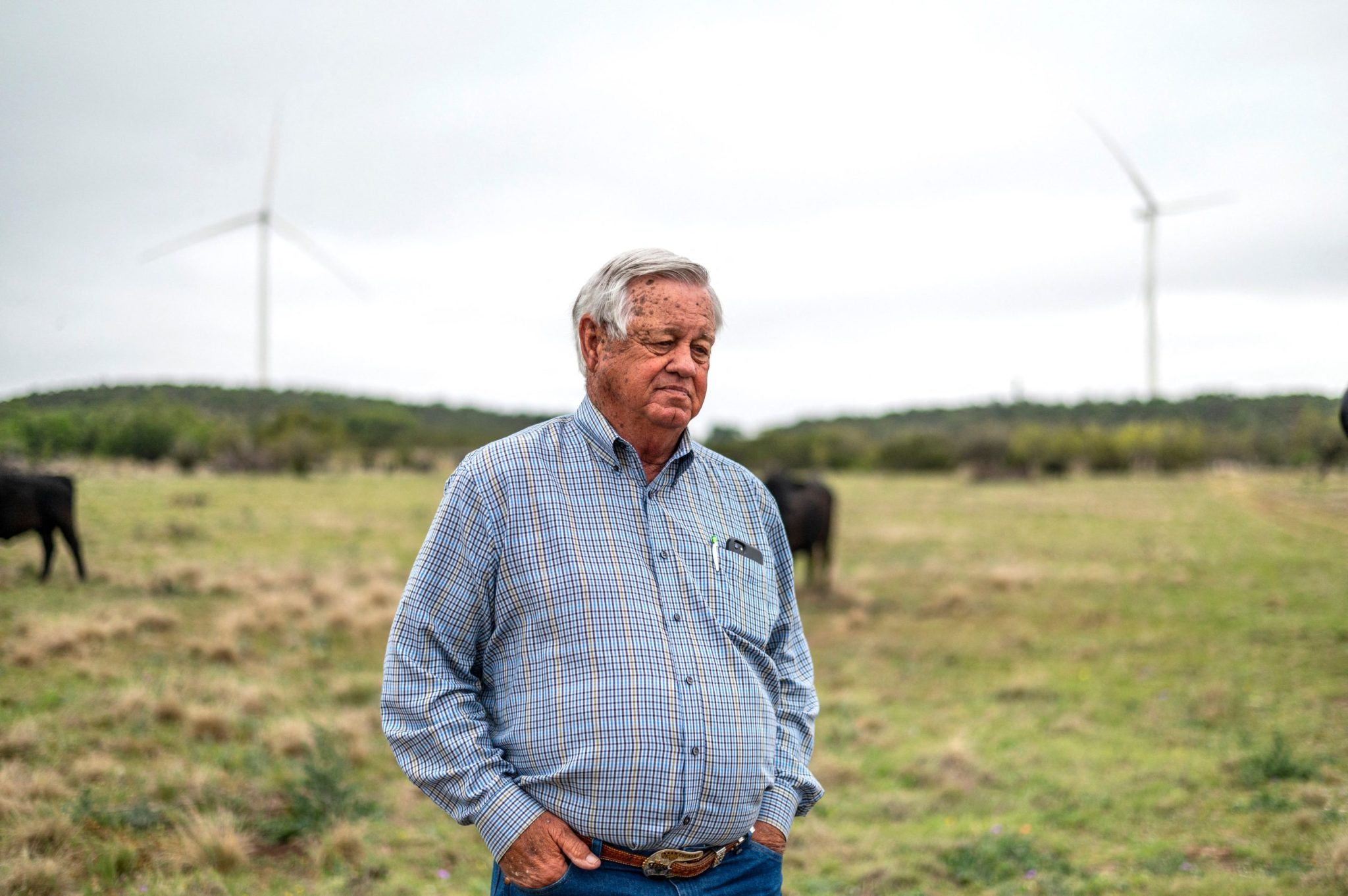 Man standing in grassy field with hands in pockets, wind turbines in the background, cloudy sky.