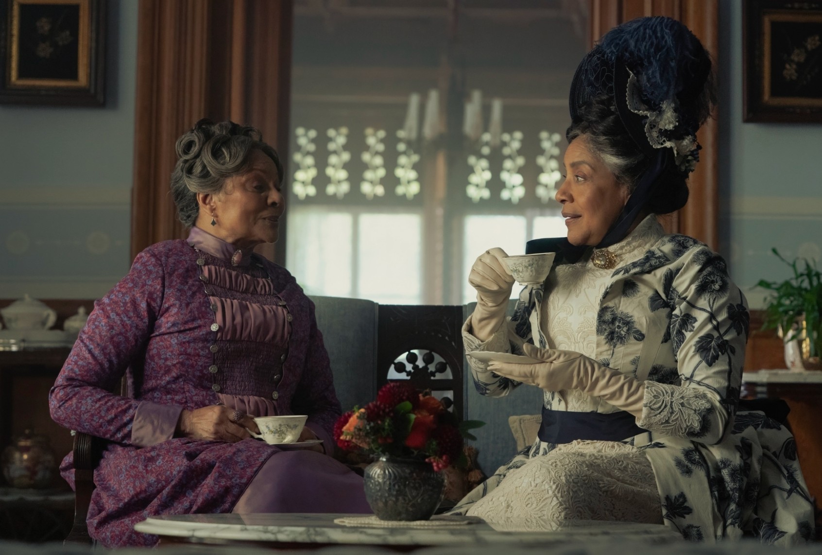 Two women in period costume enjoying tea in a vintage room, engaging in conversation with a cozy ambiance.