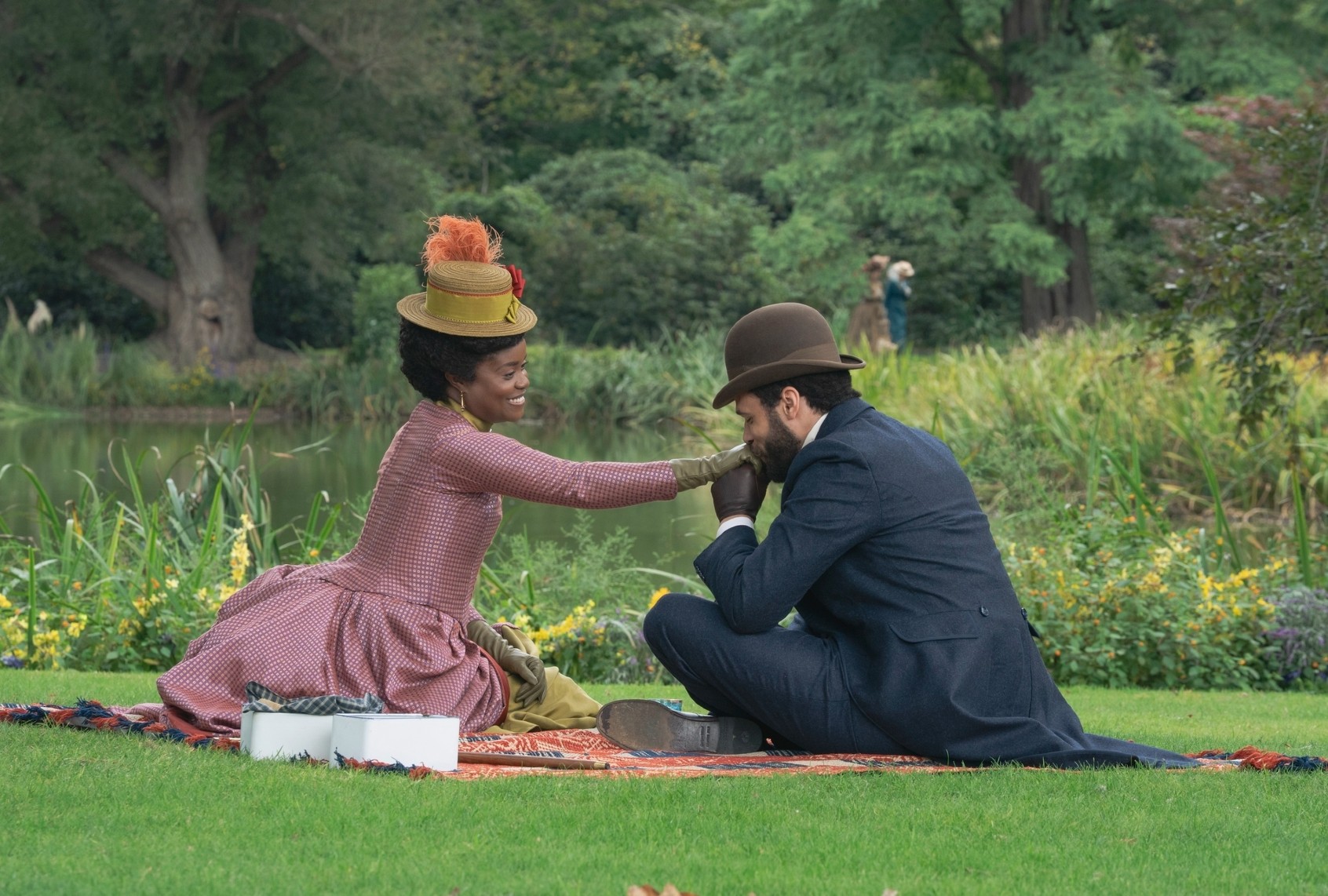 A couple in vintage attire share a joyful picnic by a pond, surrounded by lush greenery.