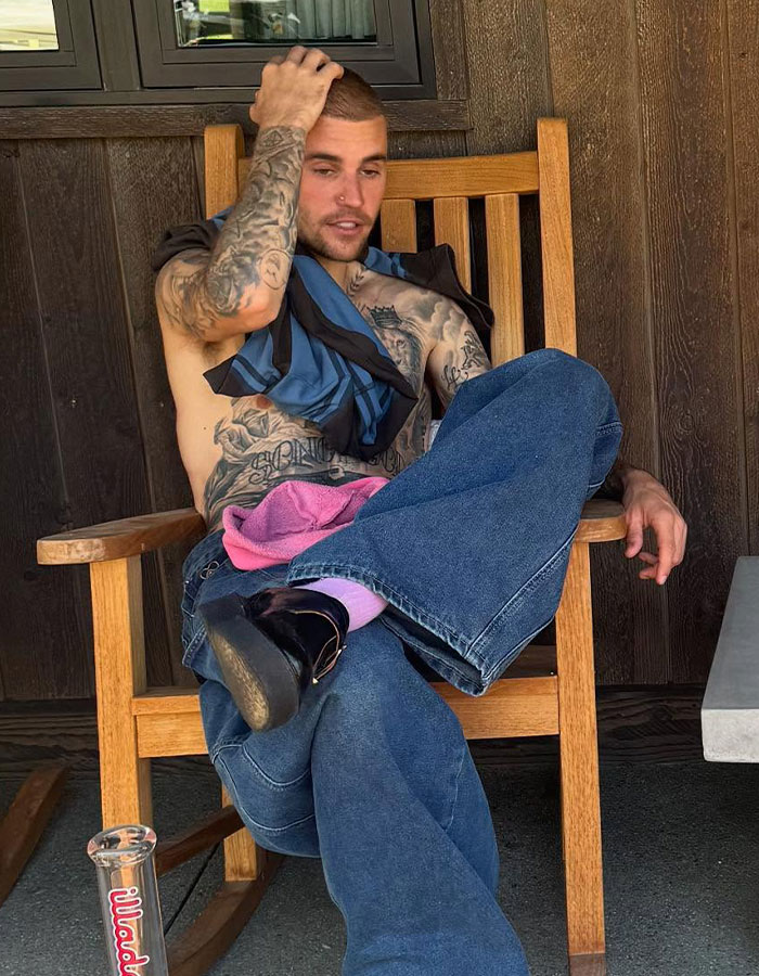 Man with tattoos sitting on a wooden rocking chair, resting his hand on his head, wearing jeans and boots.