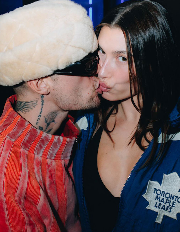 Man in sunglasses and fuzzy hat kisses woman in Toronto Maple Leafs jacket at event.