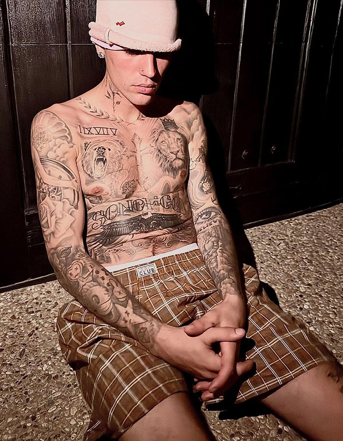 Tattooed person wearing plaid shorts and a pink hat, sitting with eyes closed against a dark background.