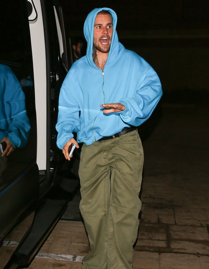 Man in blue hoodie and khaki pants exits a vehicle, holding a phone, with an expressive face.