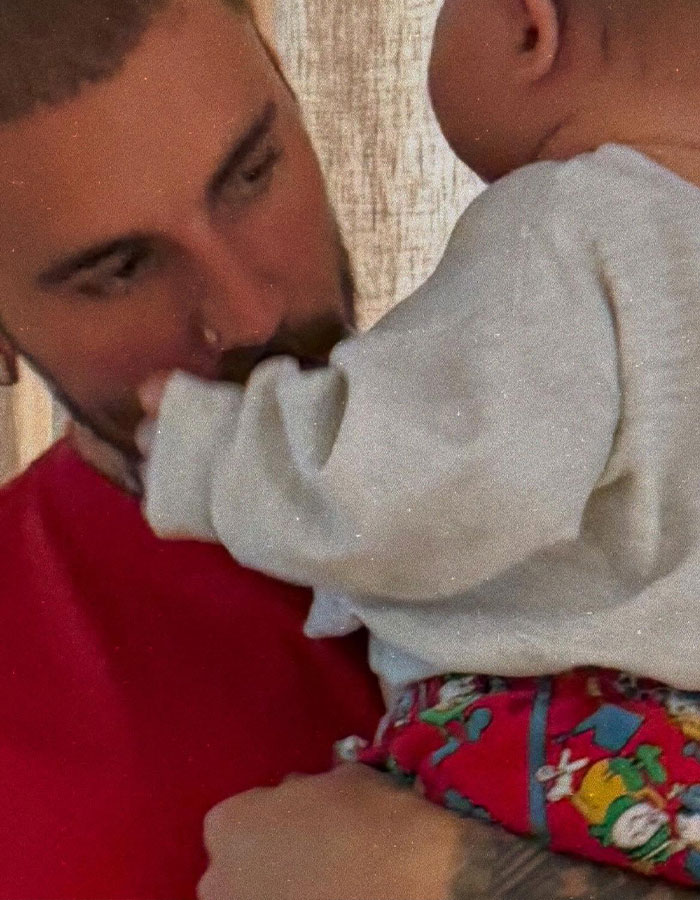 Man in red shirt gently holds and gazes at a baby wearing a colorful outfit indoors.