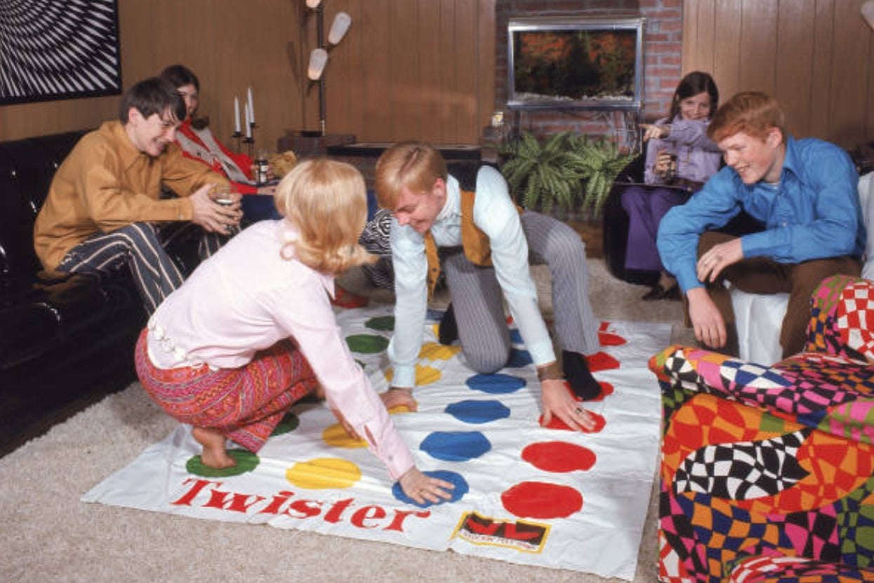 Group of friends playing Twister indoors, laughing and balancing on colorful dots, 1970s retro setting.