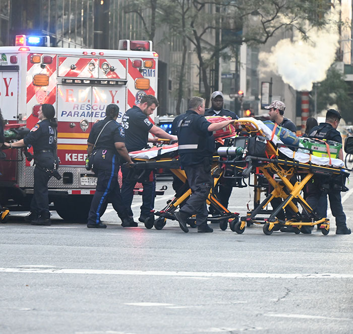 Paramedics load a patient into an ambulance in a busy city street, with a fire department vehicle and bystanders nearby.
