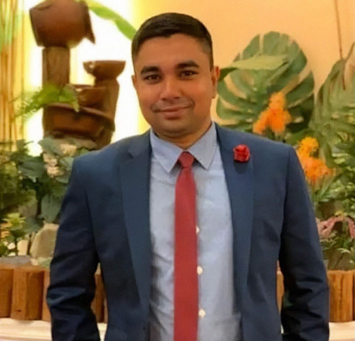 Smiling man in a blue suit and red tie stands in a garden setting with tropical plants and a wooden statue.