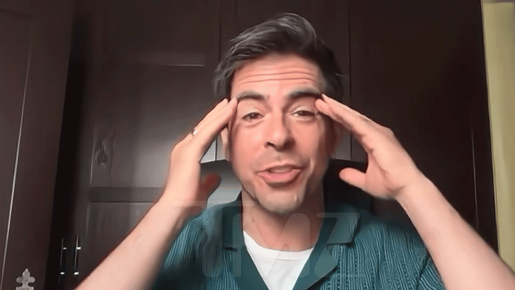 Man in green shirt talking animatedly while holding his head, standing in front of dark wooden cabinets.