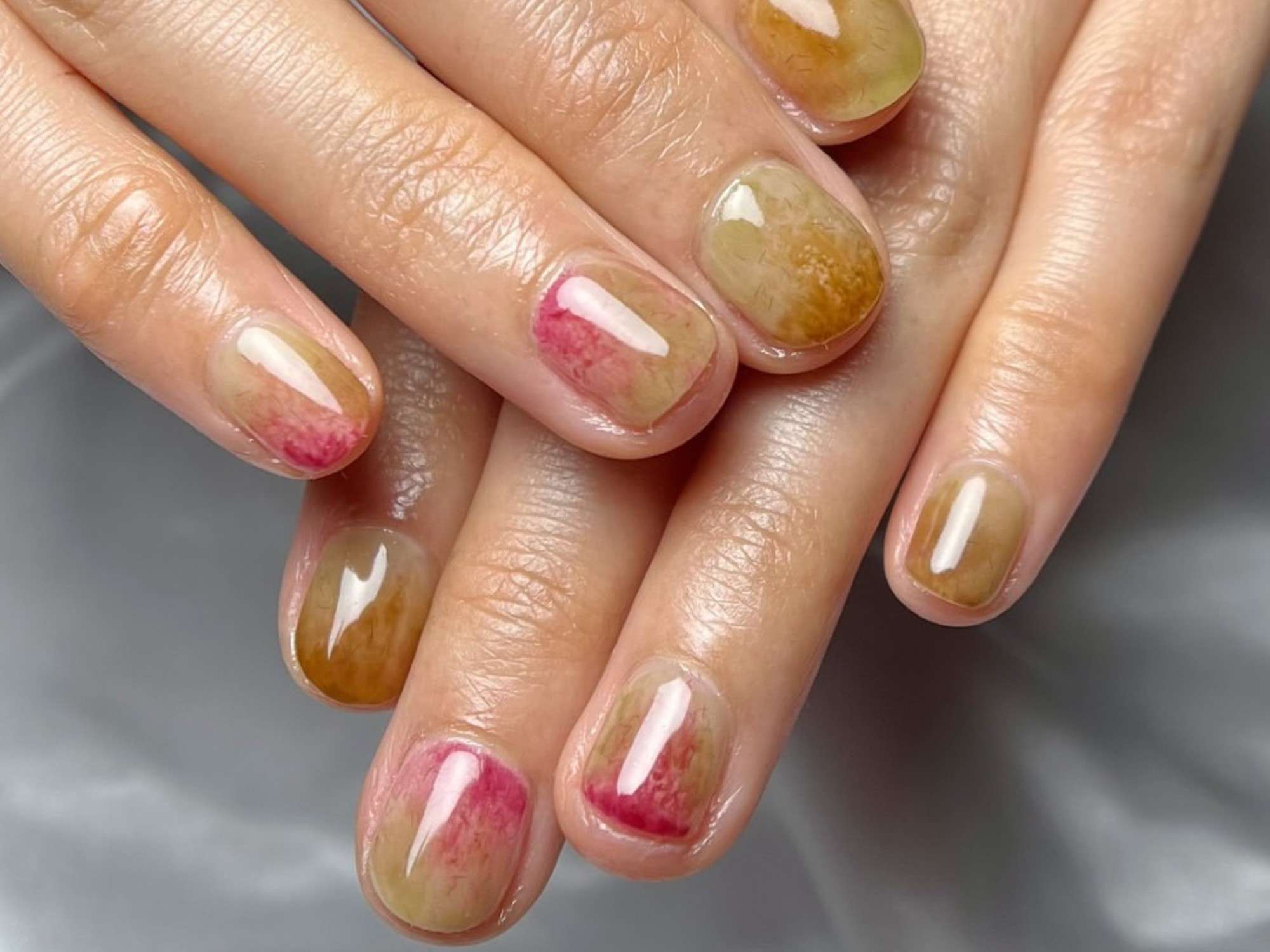 Close-up of hands featuring glossy, abstract nail art with blend of red, brown, and green hues on short, oval nails.