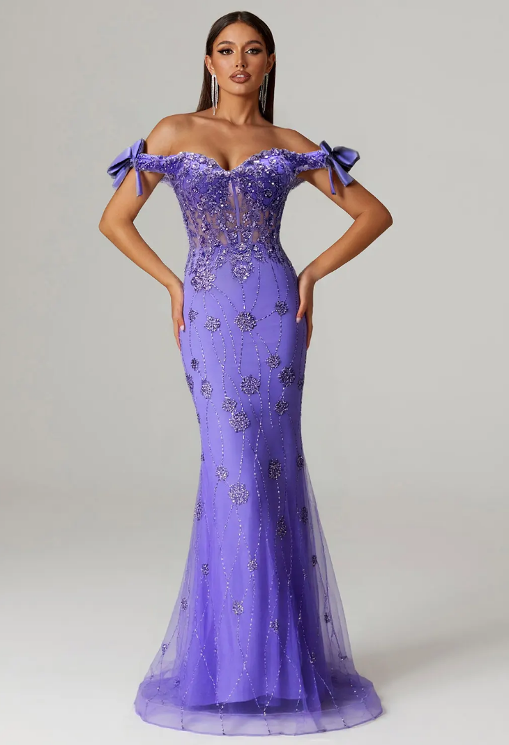 Woman in an elegant purple gown with floral embellishments and off-shoulder bows, standing against a plain background.