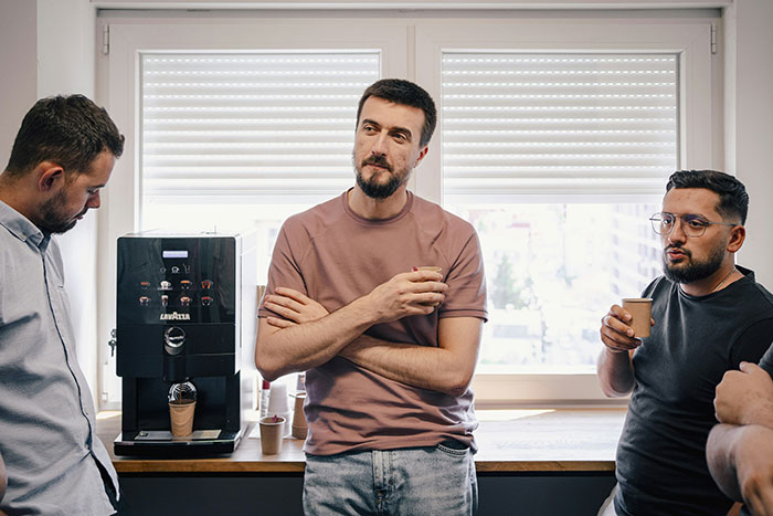 Three male co-workers standing near coffee machine, having a casual conversation in an office setting.