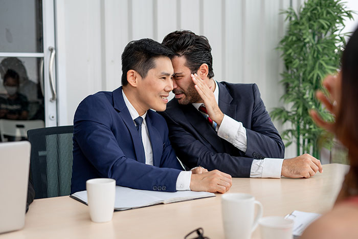Two male co-workers in suits whisper and smile during a meeting, relating to bets placed on a female colleague.