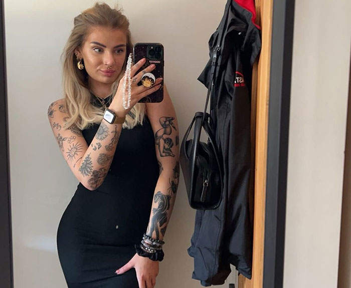Young woman with tattoos taking a mirror selfie in a black dress, highlighting male co-workers placing bets and legal action.