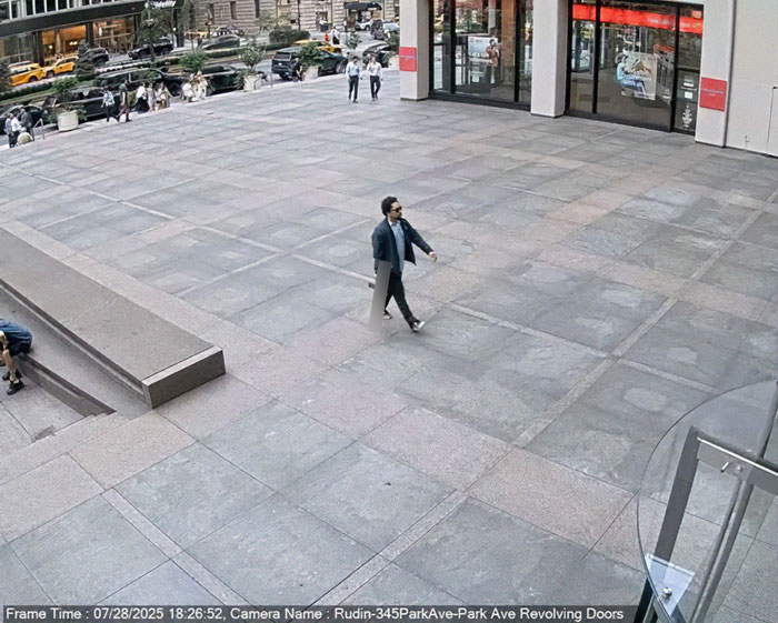 Person walking across a spacious urban plaza near revolving doors, with city buildings and traffic in the background.
