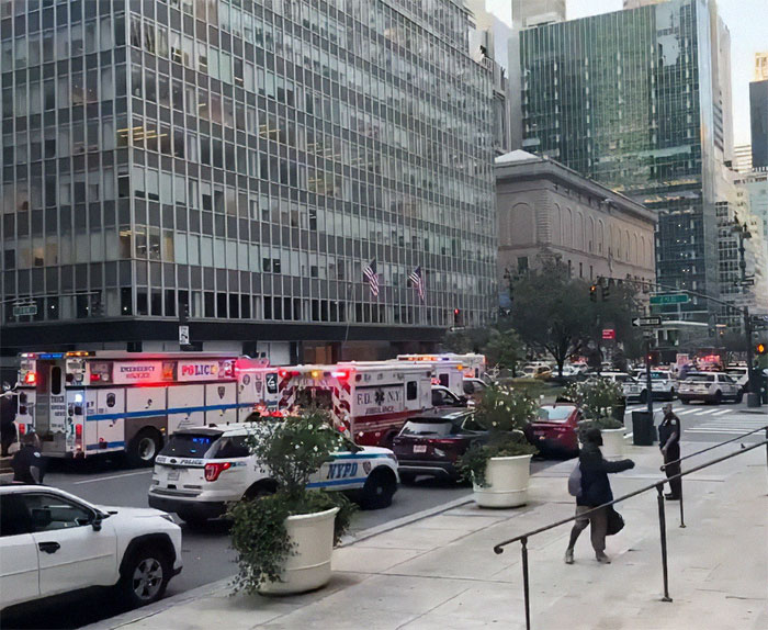 Emergency vehicles with flashing lights at a busy city intersection, surrounded by skyscrapers, pedestrians, and traffic.