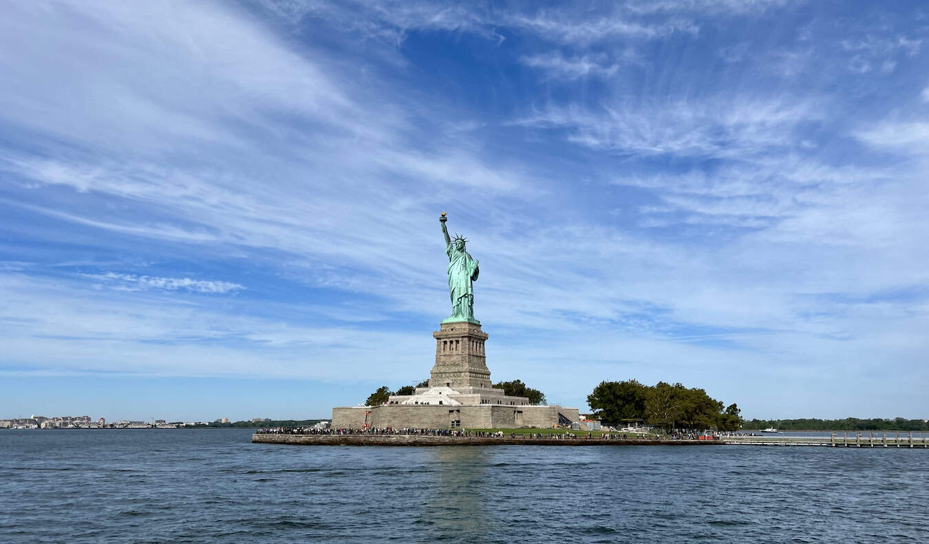 Statue of Liberty on Liberty Island under a bright blue sky, surrounded by water and distant cityscape.