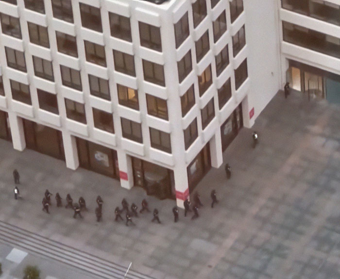 Aerial view of a group of people walking in formation by an office building on a large city plaza.
