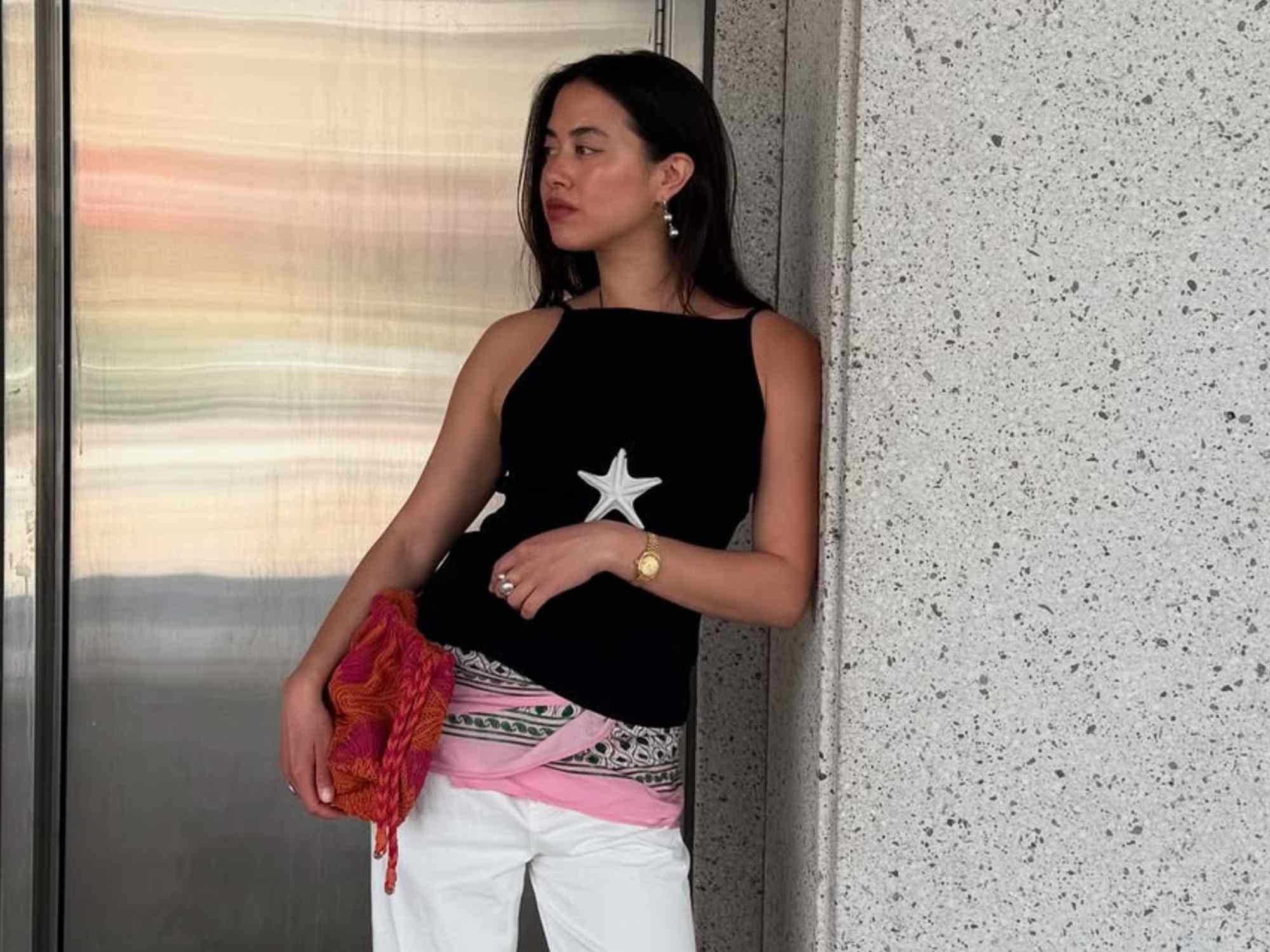 Woman in star-print tank top, holding a red bag, leans against a wall, looking away. Casual summer style.
