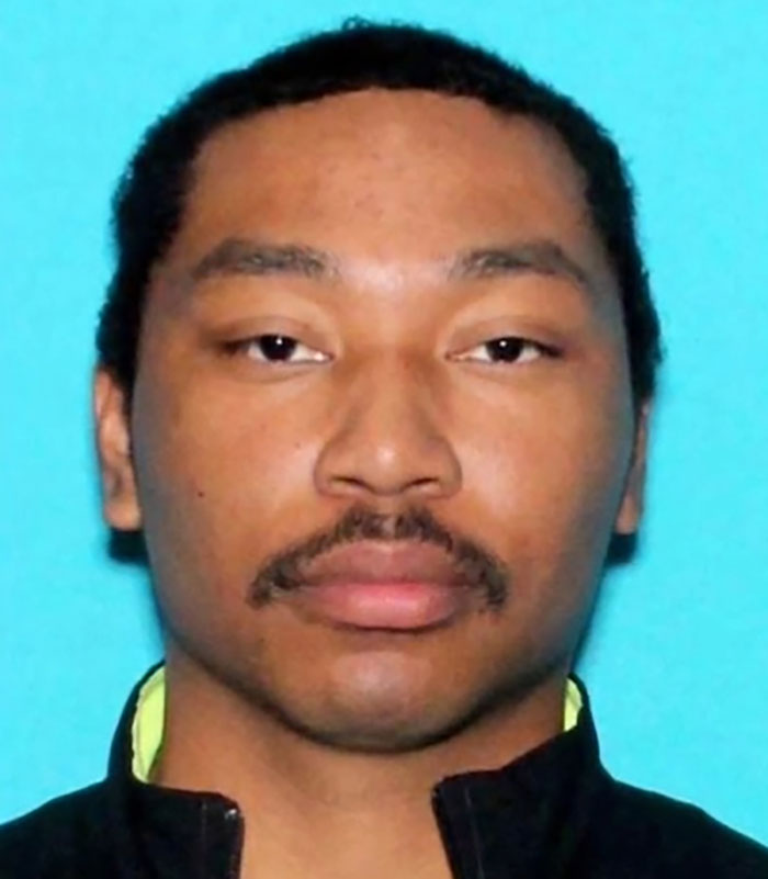 A person with short hair and a mustache, wearing a zipped jacket, looking directly at the camera against a blue background.