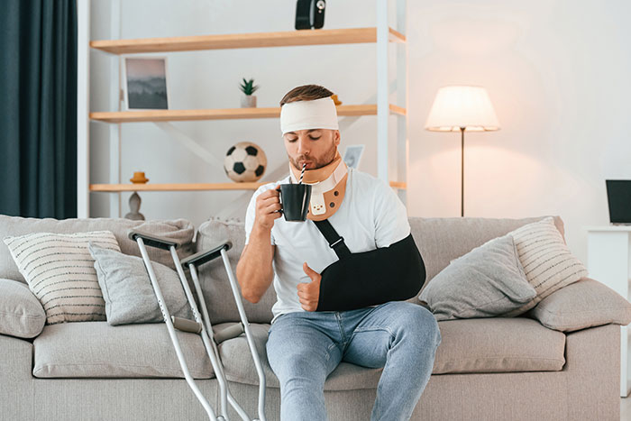 Man with head bandage and arm in sling sipping from mug, sitting on couch beside crutches, giving thumbs up.