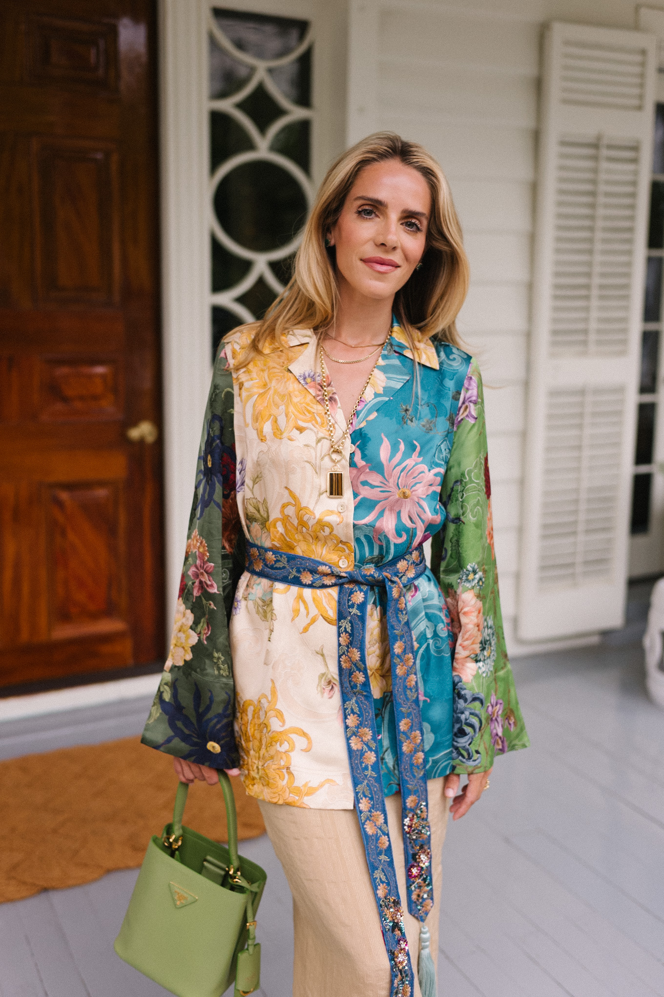 Woman in vibrant floral outfit with green handbag on porch. Elegant, stylish fashion statement.