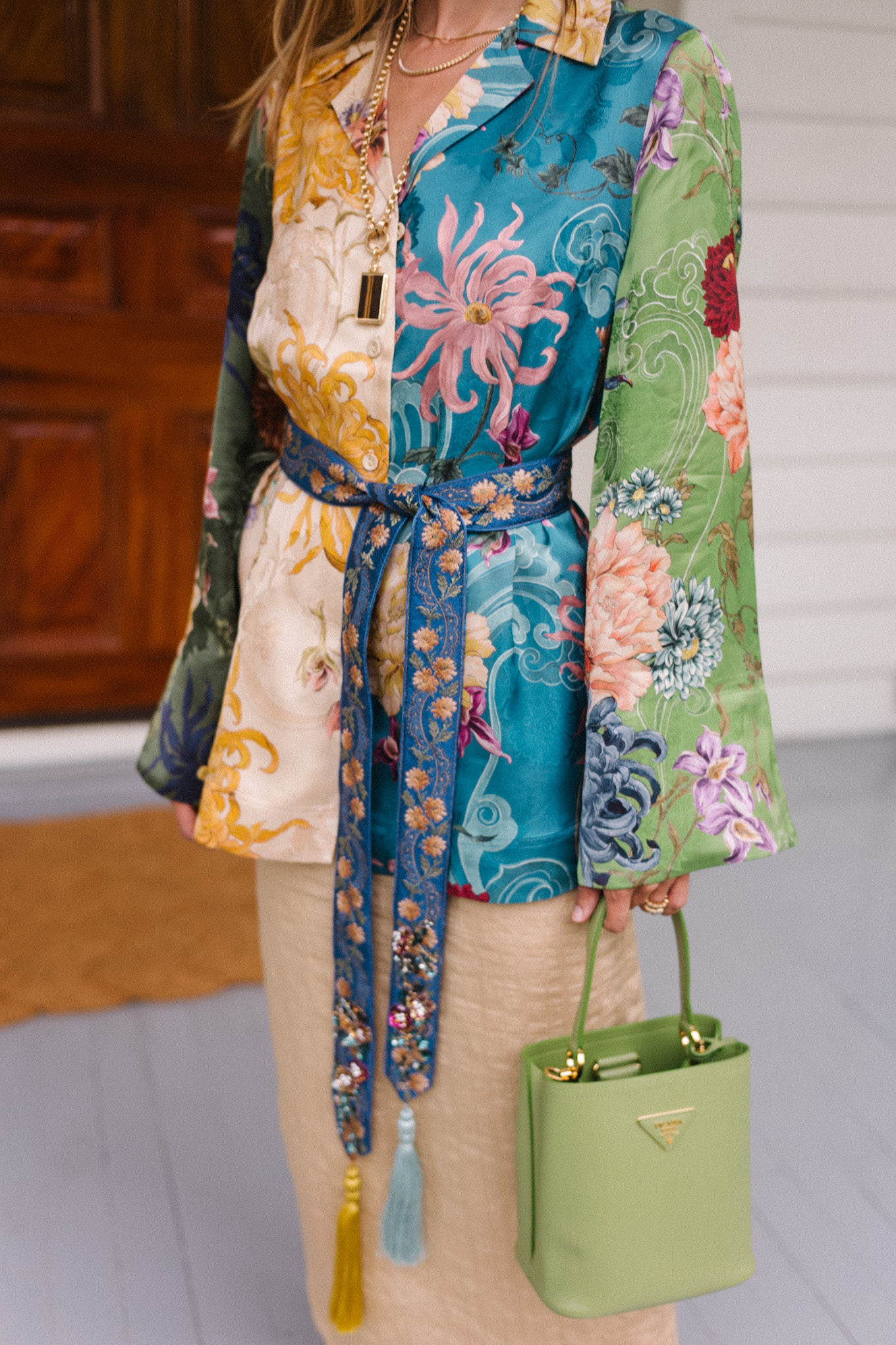 Woman in floral-patterned blouse with belted waist, holding a light green handbag, standing on a porch.