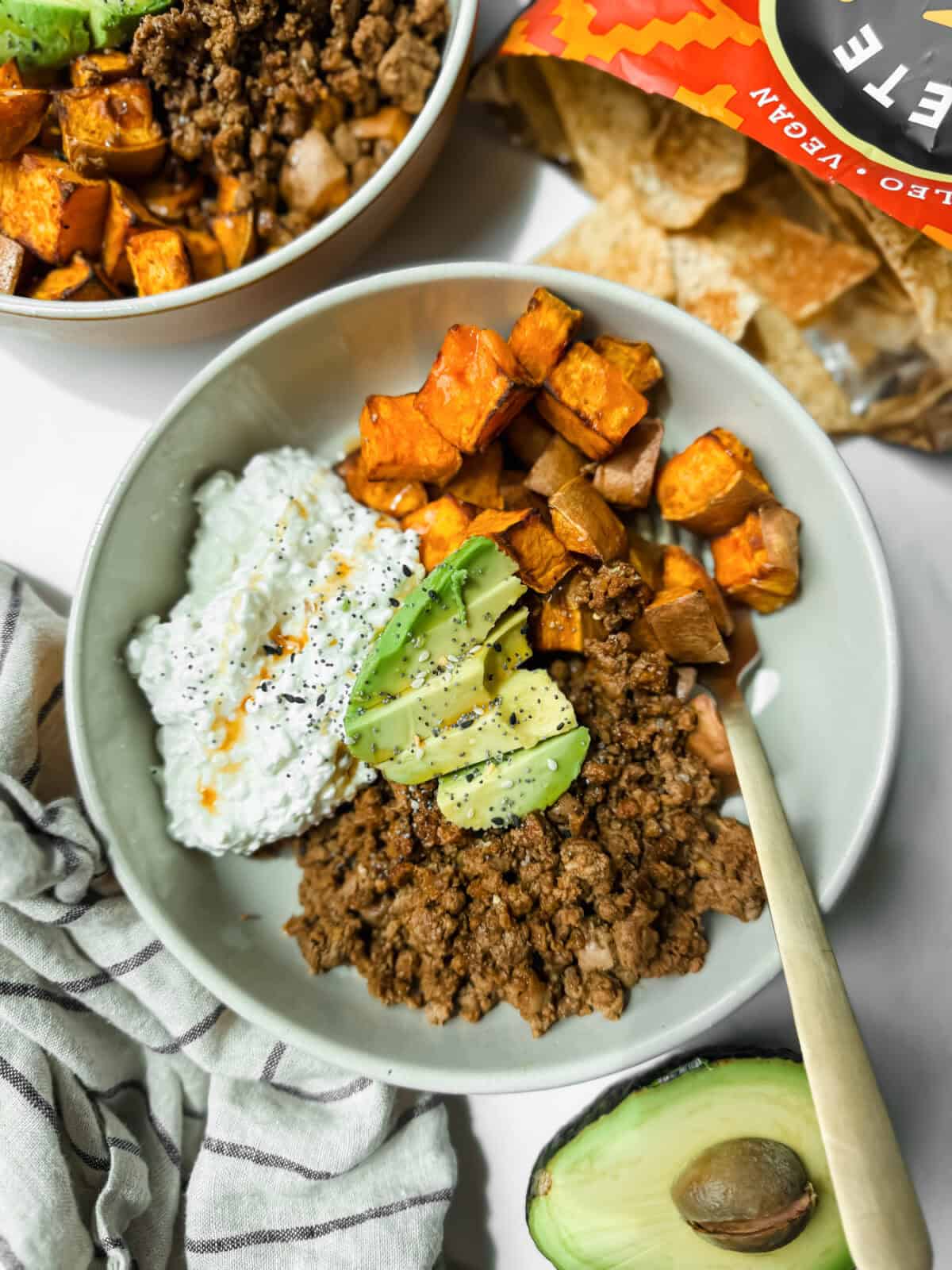 A bowl with ground beef, roasted sweet potatoes, cottage cheese, and avocado slices sits next to an open bag of tortilla chips and a halved avocado.