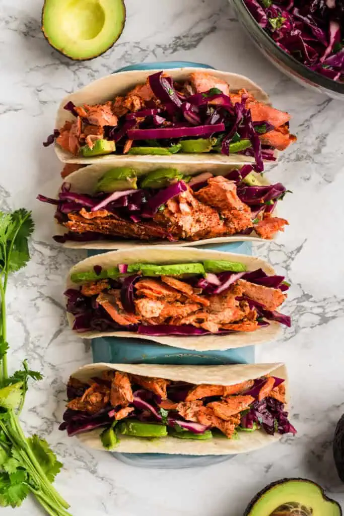 Four chipotle salmon tacos in a taco holder on a marble counter, avocado on the side.