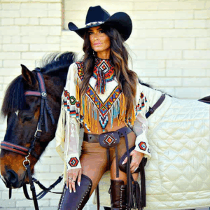 Woman in vibrant Native American-inspired attire and cowboy hat standing beside horse against brick wall.